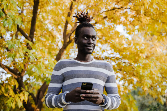 Smiling man looking away while holding smart phone standing against yellow tree in park during autumn