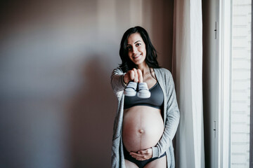 Smiling pregnant woman showing baby booties while standing against wall at home