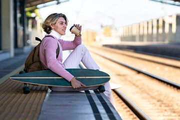 Smiling young man holding reusable cup while sitting with longboard at edge of railroad platform