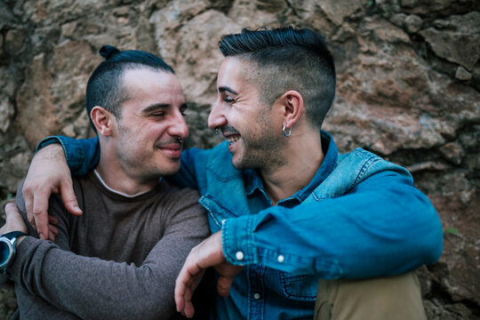 Happy gay couple smiling at each other while sitting against stone wall