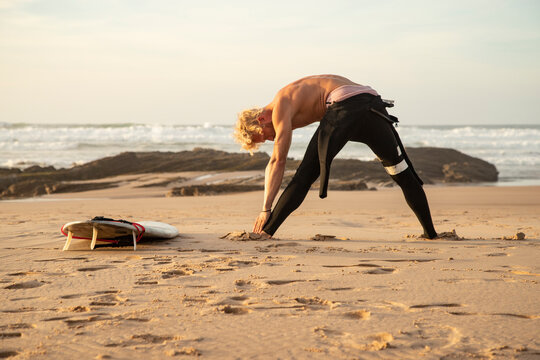 Shirtless Male Surfer Exercising By Surfboard At Beach