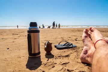 A thermos with a mate, a pair of flip flops and a pair of crossed feet resting in the sand on the beach with people walking in the background