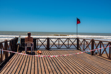 Mar del Tuyú, Buenos Aires, Argentina; December 06, 2020: Lifeguard sitting on a deck next to the tidal flag. A do not cross link because social distancing during COVID-19