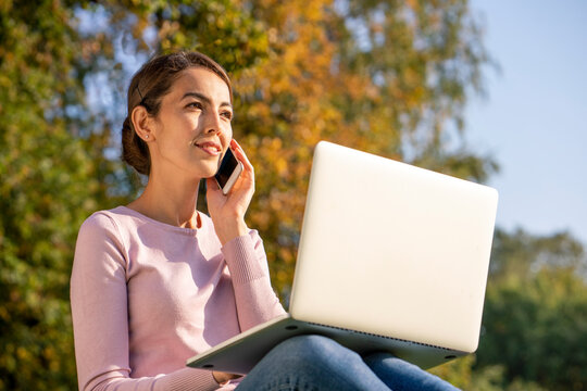 Young female entrepreneur talking on smart phone while sitting with laptop in park during autumn