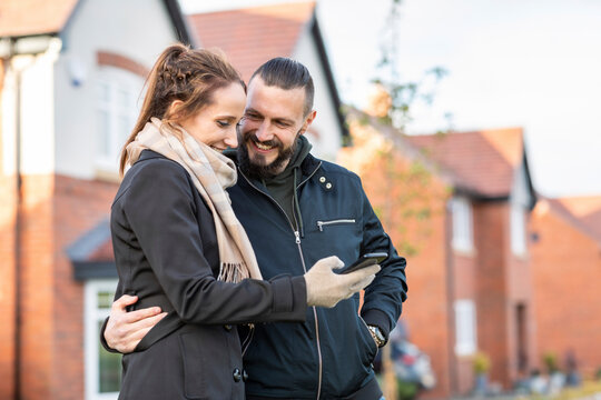 Smiling man looking at girlfriend using smart phone while standing against new houses