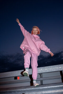 Young Woman With Pink Hair Wearing Pink Track Suit Standing On One Leg On Road Barrier At Night