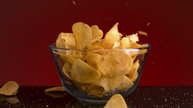 Close up of a small glass bowl and many potato chips falling into it and on the dark table. Stock footage. Spicy delicious chips on red wall background, concept of eating junk food. 