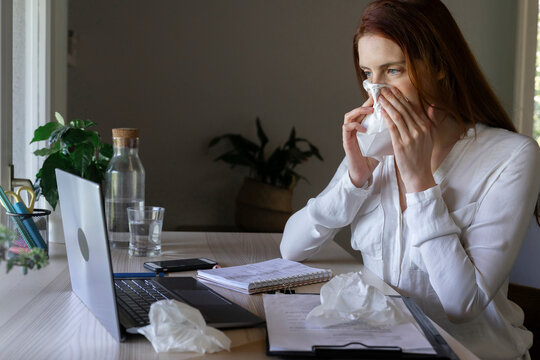 Sick woman talking to doctor on laptop at home, cleaning nose with tissue paper