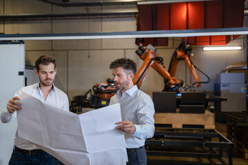 Team with blueprint having discussion while standing against robotic arm machine at factory