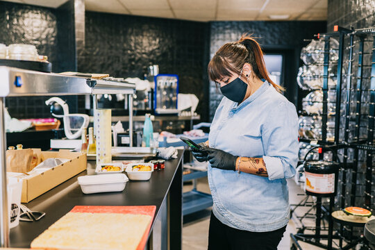 Female Chef Using Smart Phone While Standing At Kitchen Counter In Restaurant During Coronavirus