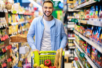 Young cheerful bearded man shopping in supermarket with trolley cart
