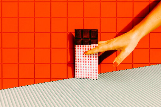 Woman Hand Holding Dark Chocolate Bar Against Red Tile Wall