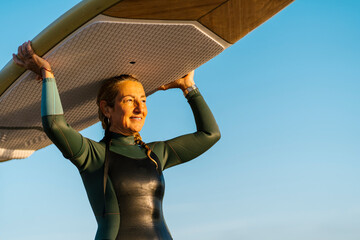 Mature woman carrying paddleboard on head while standing against blue sky