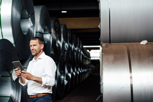 Businessman holding digital table while standing by steel rolls at industry
