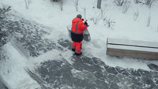 Janitor in bright orange workwear shoveling snow on the paths in the park.