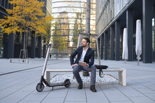 Entrepreneur with digital tablet sitting by e-scooter on bench in city