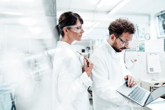 Female scientist standing by male colleague using laptop at laboratory