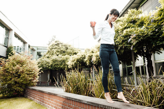 Smiling carefree businesswoman holding coffee cup while walking on retaining wall at industry