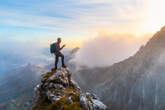 Pensive Hiker Using Smart Phone On Mountain Peak During Sunrise At Bergamasque Alps, Italy