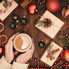 Christmas and New Year rustic traditional composition. Hot coffee cup in female hanads. Festively decorated cedar branches on dark brown old wooden board. Gift boxes, balls, stars and cane lollypops.