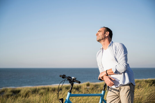 Man with eyes closed leaning on bicycle at beach against clear sky