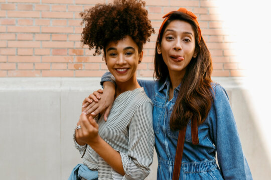 Young woman sticking tongue out while standing with friend against wall