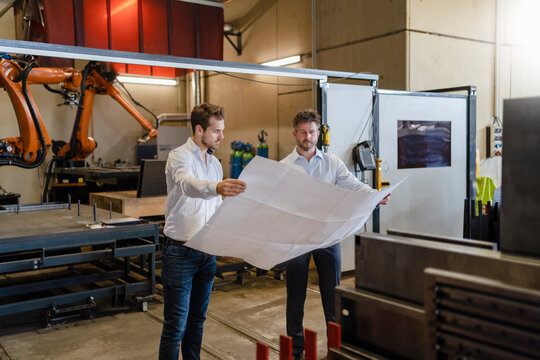 Businessmen studying blueprint of business while standing against automated machine at factory - Powered by Adobe