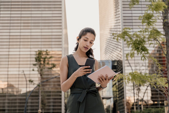 Businesswoman holding note pad using smart phone while standing against modern buildings in city