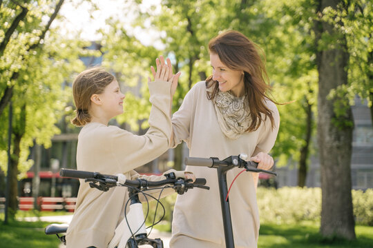 Happy mother and daughter giving high five while standing against trees in city park