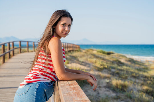 Beautiful teenage girl standing on boardwalk at beach against clear blue sky