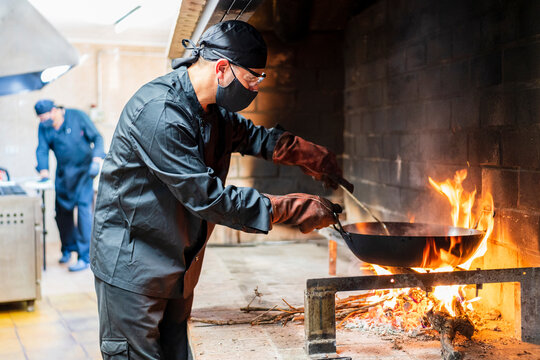 Traditional cooking of paella in restaurant kitchen, chef wearing protective mask