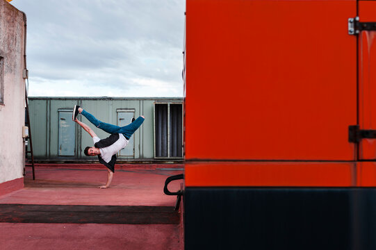Stylish bboy performing a break dance stance close to red and green machinery in the rooftop of a building 