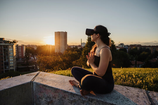 Relaxed woman using VR glasses while performing yoga in prayer positon on retaining wall in city