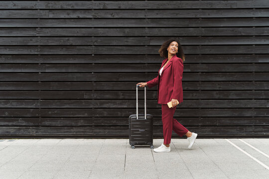 Young businesswoman looking back while walking with luggage on footpath by black wooden wall in city