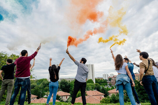 Male and female friends playing with smoke bombs against cloudy sky in park