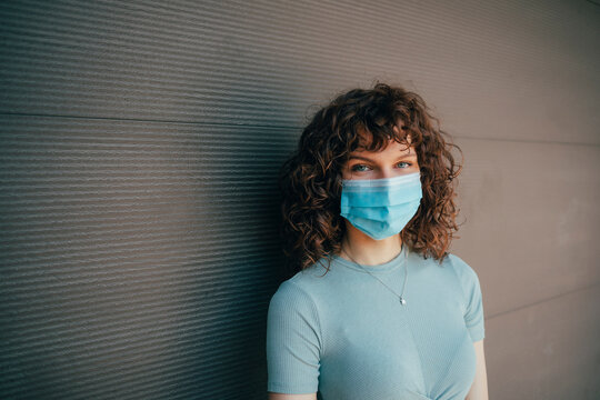 Brunette curly woman wearing protective mask in front of grey wall