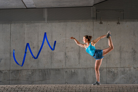 Young woman spinning ribbon standing on one leg against concrete wall under bridge