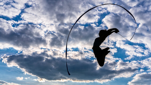 Young woman holding ribbon jumping while doing aerobics against cloudy sky