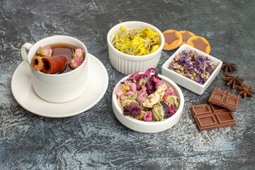 sidewise view of a cup of herbal tea with bowls of dry flowers and sweets on grey ground