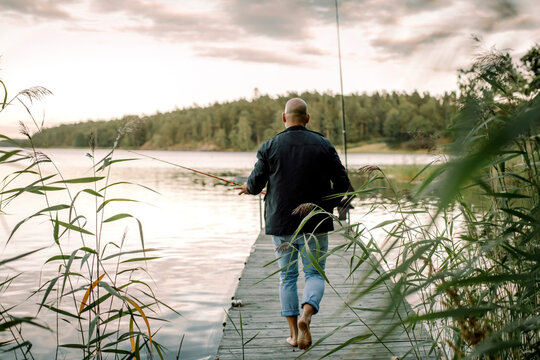 Fototapeta Rear view of man with fishing rod walking on pier against lake during sunset