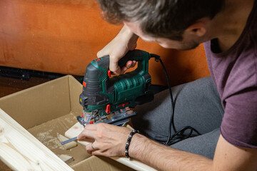 man using electric jigsaw at home to cut piece of wood board on a cardboard box. Do it yourself.