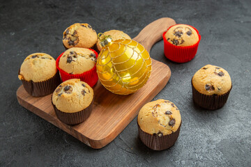 Front close view of freshly baked delicious small cupcakes with chocolates and accessory on wooden board on the right side of dark background