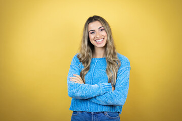 Pretty blonde woman with long hair standing over yellow background with a confident smile showing teeth with arms crossed