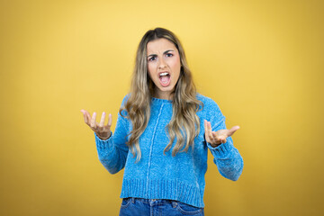 Pretty blonde woman with long hair standing over yellow background crazy and mad shouting and yelling with aggressive expression and arms raised. Frustration concept.