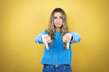 Pretty blonde woman with long hair standing over yellow background doing the negative sign showing dislike with thumb down