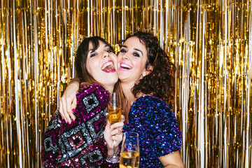 Two women in elegant clothes and Christmas decorations behind them holding glasses of champagne while hugging each other celebrating a New Year's party together. New Year's Eve at home concept