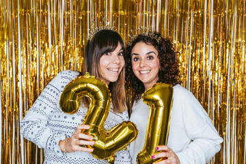 Two brunette women in pajamas and Christmas headbands smiling at the camera and holding golden balloons with the number 2021 in a New Year's party at home. 2020 New Year's Eve concept