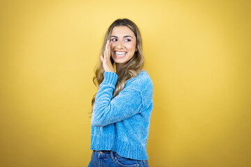 Pretty blonde woman with long hair standing over yellow background with hand on mouth telling secret rumor, whispering malicious talk conversation