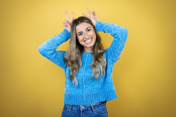 Pretty blonde woman with long hair standing over yellow background Posing funny and crazy with fingers on head as bunny ears, smiling cheerful