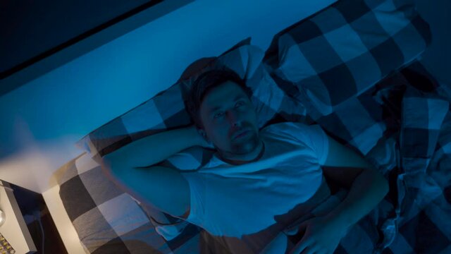 Stressed man trying to sleep in bed at night. Young male with open eyes suffers from insomnia and sleep disorders. Things dont feel same anymore. Blue tones. Sleepless man. Guy dont sleep at night